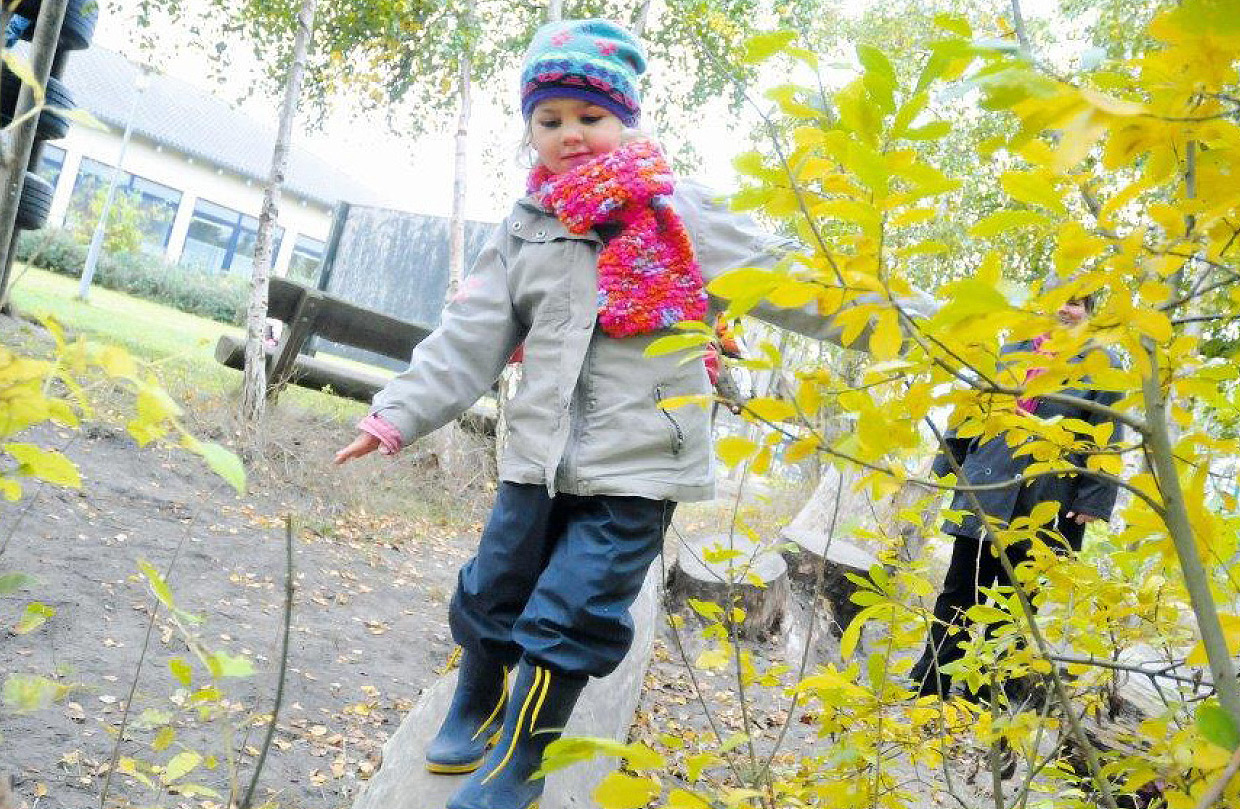 Ein Kind mit einer bunten Mütze und einem rot-pinken Schal balanciert auf einem Baumstamm im Außenbereich der Ostseeklinik Grömitz. Im Hintergrund sind Bäume und ein Gebäude der Klinik zu sehen.