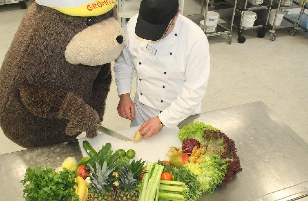Grömel, das Maskottchen von Grömitz und der Ostseeklinik mit braunem Fell und gelber Kappe, steht gemeinsam mit einem Koch in der Küche der Klinik. Grömel schneidet Gemüse. Vor den beiden liegt Gemüse, Obst und Salat auf dem Küchentisch aus Metall.