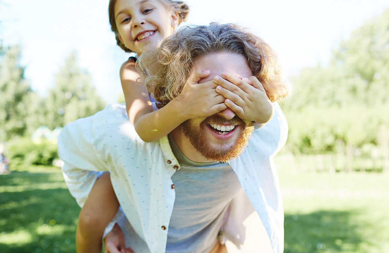 Ein lachender Vater trägt sein Kind auf seinem Rücken Schultern, wobei das Kind ihm spielerisch die Augen zuhält. Vater und Kind befinden sich im Freien, umgeben von grüner Natur bei sonnigem Wetter.