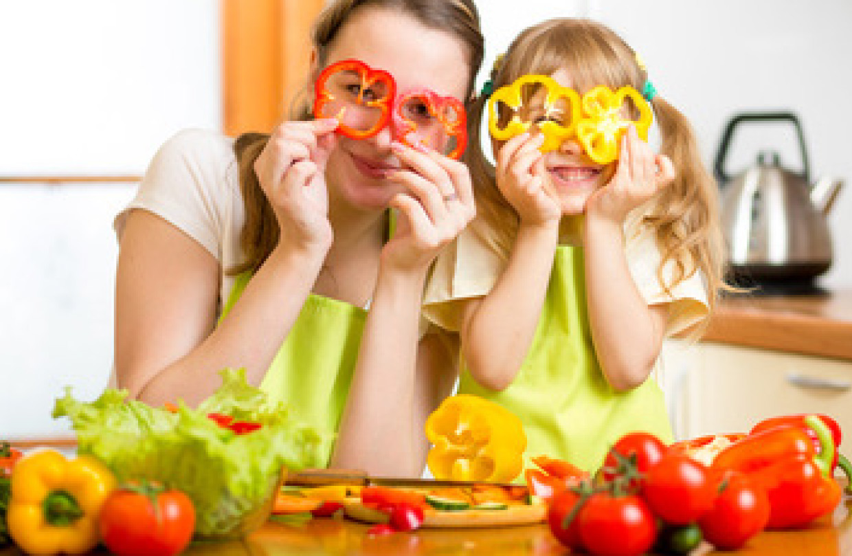  Eine lächelnde Mutter und ihre kleine Tochter halten beide Scheiben von roter und gelber Paprika vor ihre Augen wie Brillen. Sie stehen in einer hellen Küche mit Gemüse und einem Schneidebrett auf der Arbeitsfläche.