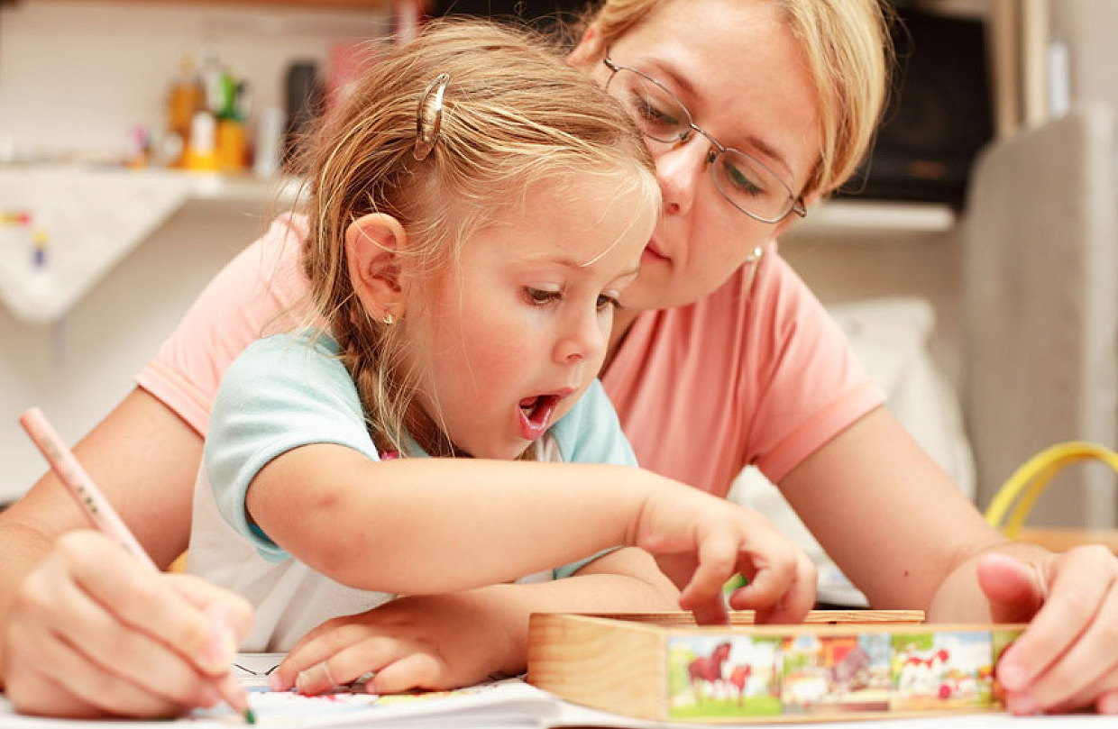 Eine Mutter mit Brille beugt sich über ihre Tochter und schreibt mit einem Bleistift auf ein Papier, während das Kind erstaunt in eine Holzkiste auf dem Tisch greift.