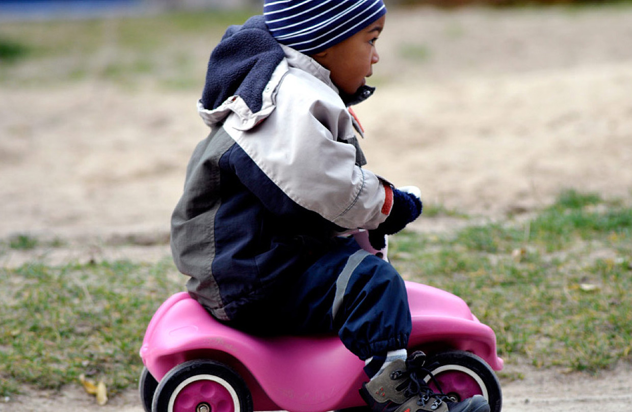 Ein kleines Kind mit Mütze, Winterjacke und Hose sitzt auf einem pinkfarbenen Bobbycar und fährt auf einem sandigen Untergrund mit Gras.