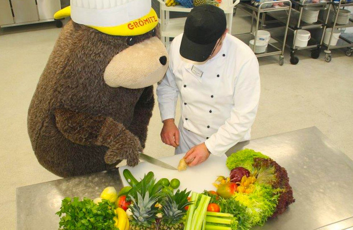 Grömel, das große braune Stofftier-Maskottchen mit gelber Kappe, und ein Koch in der Küche der Ostseeklinik Grömitz. Grömel schneidet Gemüse auf einem Küchentisch aus Metall, auf dem Gemüse, Salat und Obst liegt.