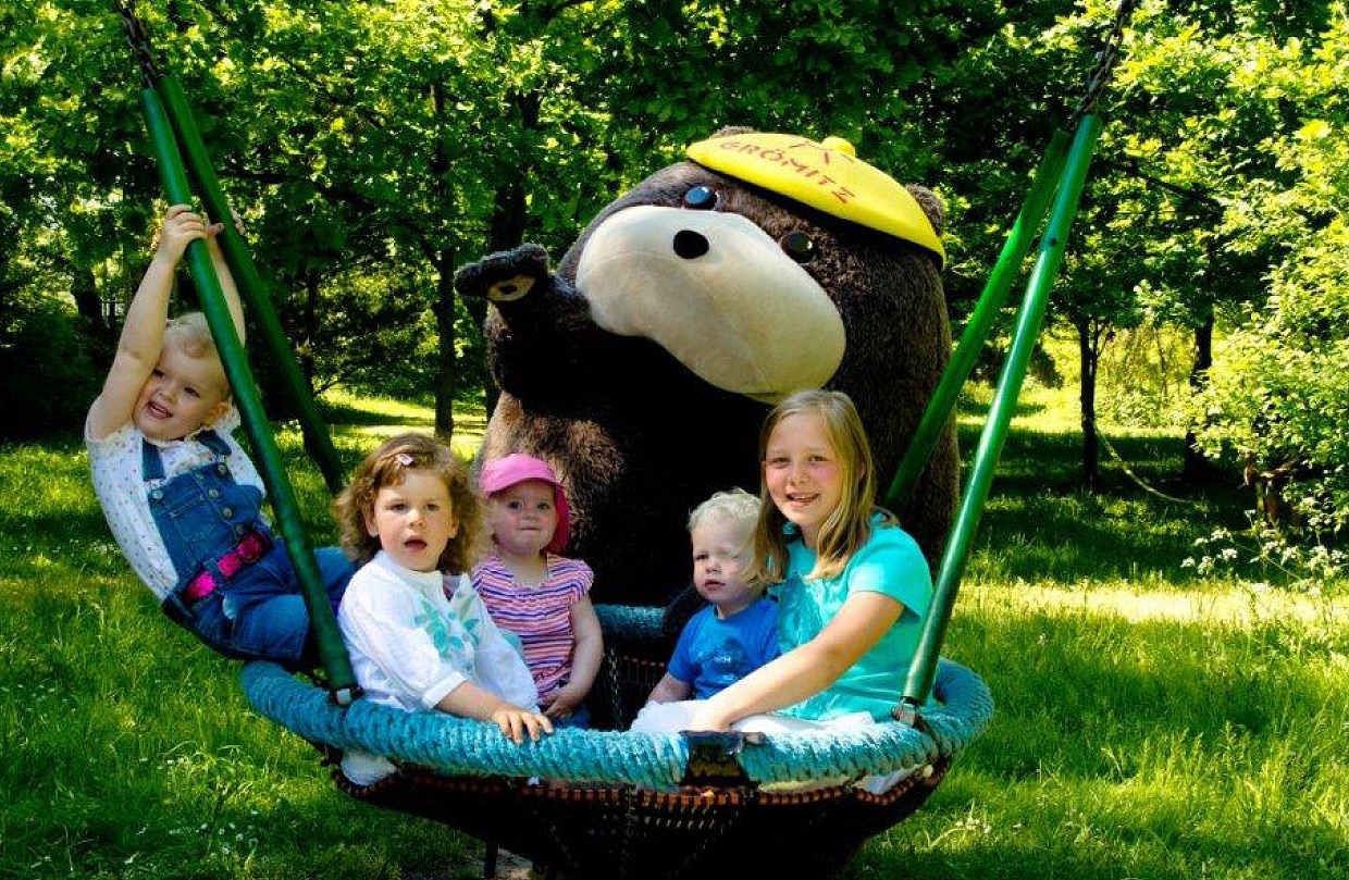 Mehrere Kinder sitzen in einer Nestschaukel im Grünen der Ostseeklinik. Die Kinder lachen und genießen den Sommertag. Grömel, das große braune Stofftier-Maskottchen mit gelber Kappe, steht hinter der Schaukel und winkt.