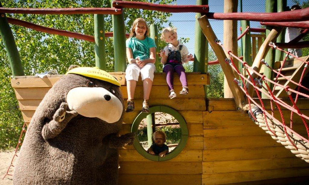 Maskottchen Grömel winkt vor einem hölzernen Spielschiff auf dem Spielplatz der Ostseeklinik Grömitz; zwei Kinder sitzen auf dem Schiff, ein weiteres schaut aus einem Bullauge.