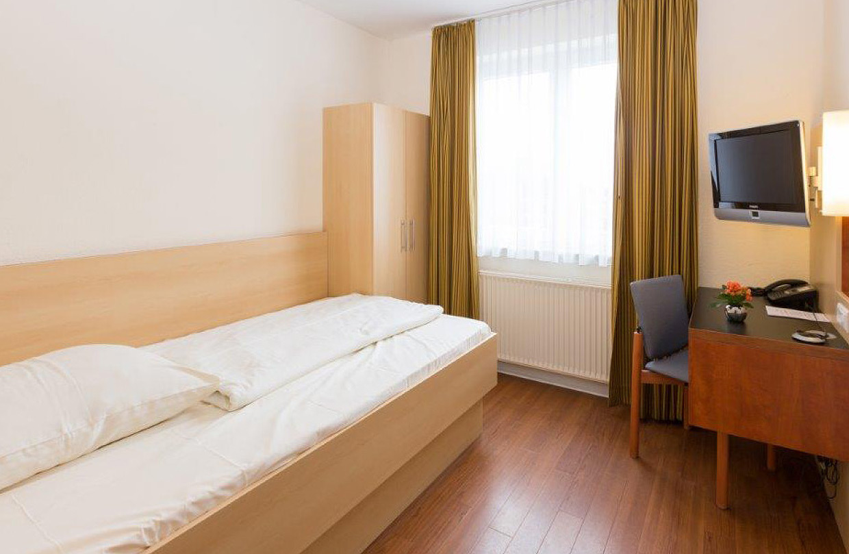 Ein helles und freundlich eingerichtetes Patientenzimmer in der Klinik. Zu sehen sind ein Bett links, ein Fenster vor Kopf und rechts ein Schreibtisch mit Telefon und Stuhl, darüber ein Fernseher an der Wand.