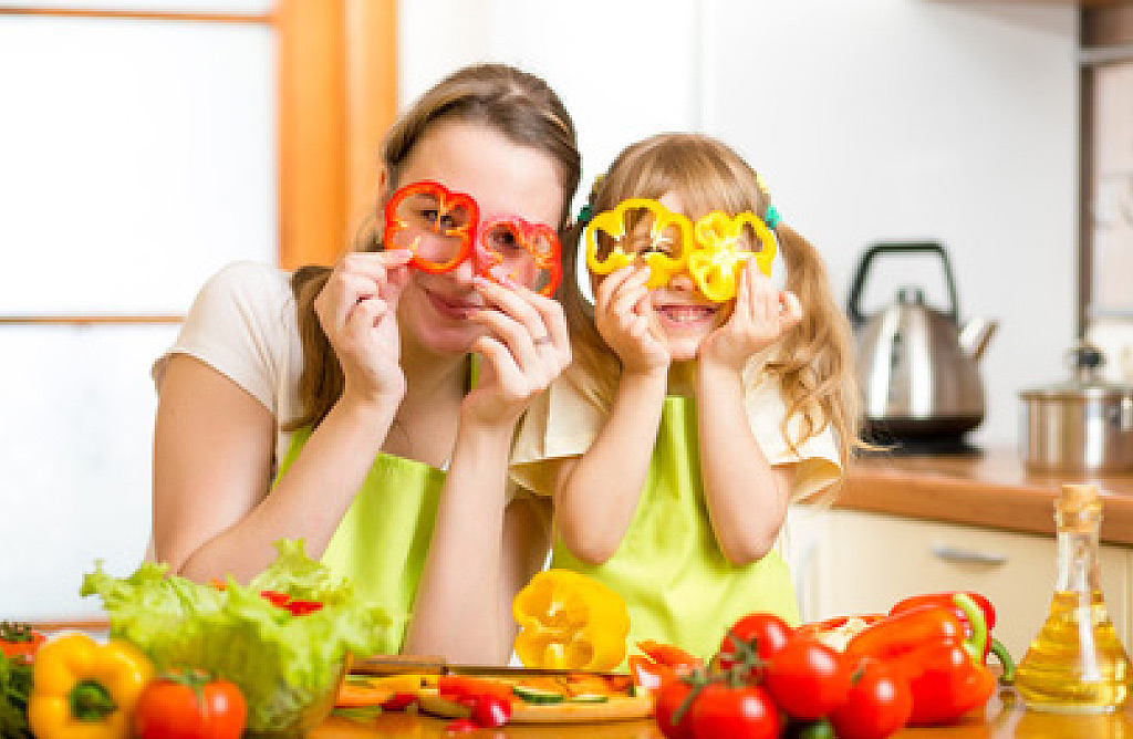 Eine lächelnde Mutter und ihre kleine Tochter halten Scheiben von roter und gelber Paprika vor ihre Augen wie Brillen. Sie stehen in einer hellen Küche mit Gemüse und einem Schneidebrett auf der Arbeitsfläche.