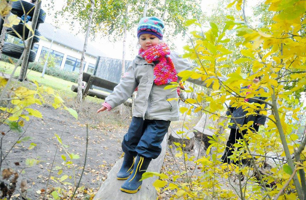 Ein Kind mit einer bunten Mütze und einem rot-pinken Schal balanciert auf einem Baumstamm im Außenbereich der Ostseeklinik Grömitz. Im Hintergrund sind Bäume und ein Gebäude der Klinik zu sehen.