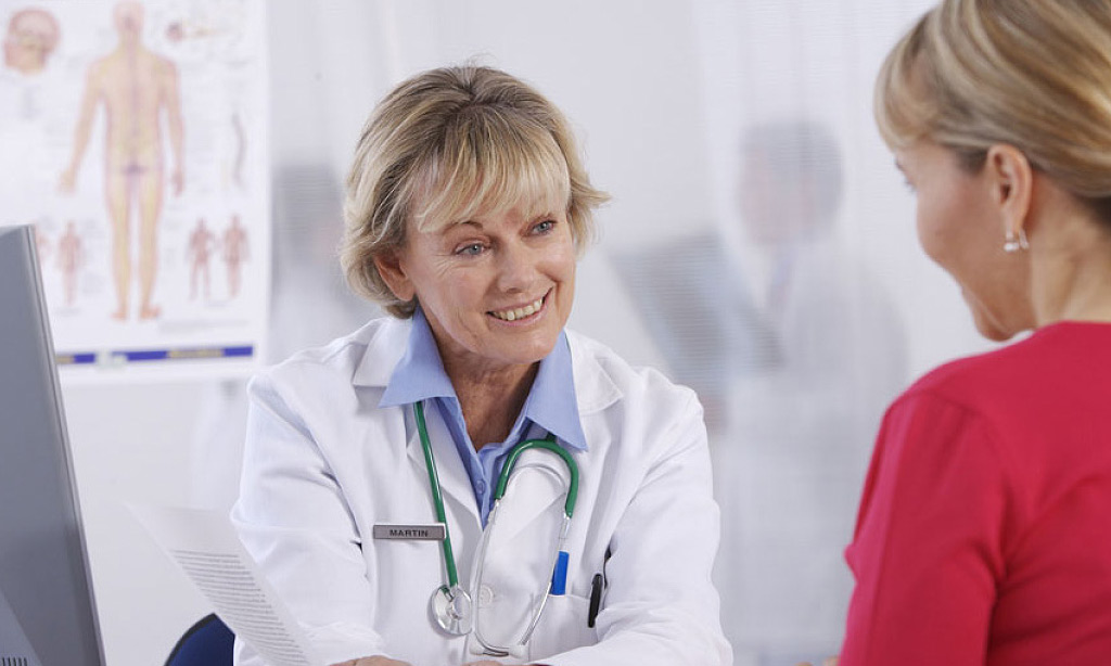 Eine Ärztin mit blonden Haaren weißem Arztkittel lächelt eine Patientin an, die ihr gegenüber sitzt. Im Hintergrund ist ein anatomisches Poster zu sehen.