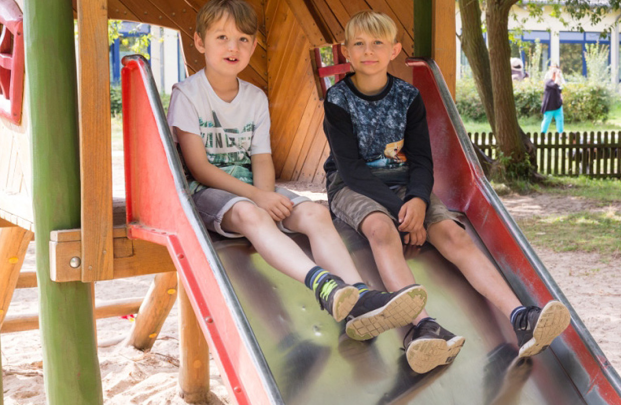 Zwei Jungen sitzen auf einer Holzrutsche auf dem Spielplatz im Außenbereich der Klinik, im Hintergrund sind ein Zaun, Bäume und ein Klinikgebäude zu sehen.