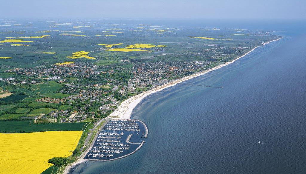 Luftaufnahme des Küstenortes Grömitz an der Ostsee mit Strand, Promenade, Hafen und gelb blühenden Feldern im Hinterland unter blauem Himmel. Rechts ist die ruhige Ostsee zu sehen.