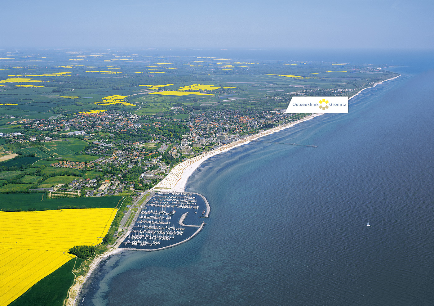 Luftaufnahme der Küste von Grömitz mit dem Ostseestrand, einem Hafen und der Stadt. Im Hinterland sind grüne Felder und gelbe Rapsfelder zu sehen. Die Ostseeklinik ist in Strandnähe markiert.