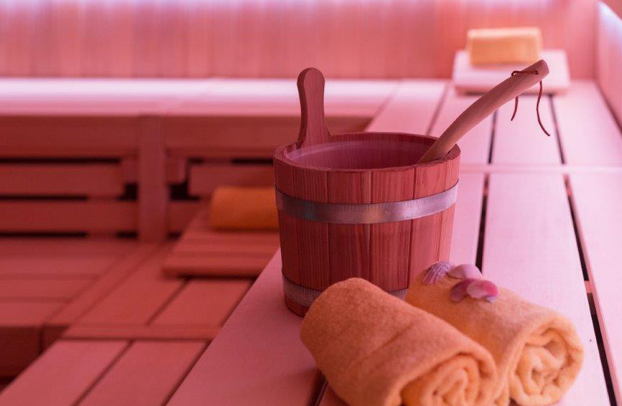 Der Innenraum einer finnischen Sauna mit Holzverkleidung, mehrstufigen Sitzbänken, einem hölzernen Aufgusseimer mit Kelle und gerollten Handtüchern davor.