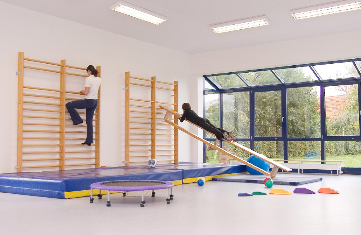 Zwei Kinder in einer hellen Sporthalle machen Übung auf einer Holzbank und an einer Sprossenwand. Weitere Sportgeräte wie ein Trampolin, Bälle und Matten sind zu sehen.