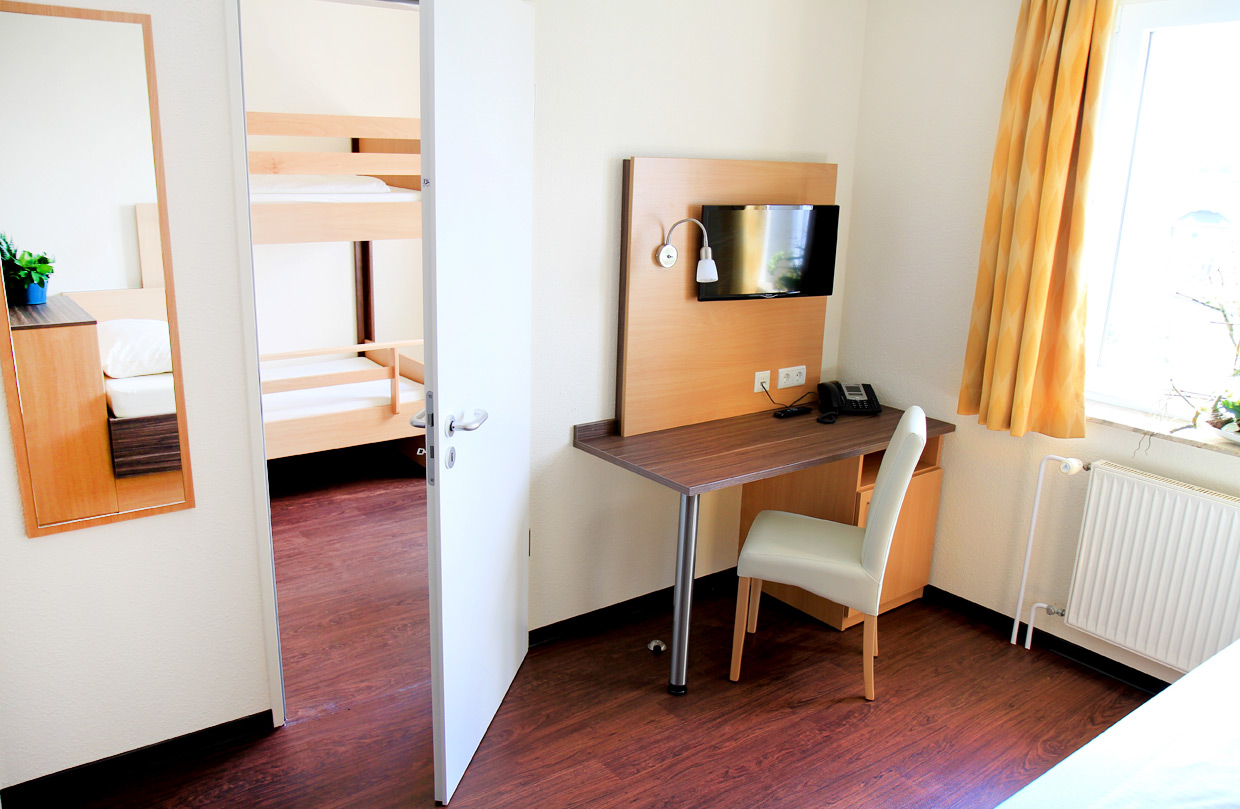 Helles Patienten-Appartement mit Bett, Schreibtisch mit Telefon und gepolstertem Stuhl davor, darüber ein Wand-Fernseher. Durch eine offene Tür im ist ein Etagenbett für Kinder zu sehen.