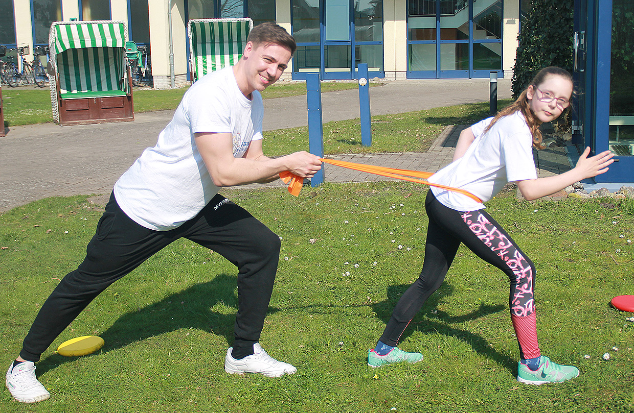 Ein Mädchen und ein Kinderbetreuer führen auf dem Gelände der Ostseeklinik Grömitz eine sportliche Bewegungsübung im Freien durch, wobei der Betreuer das Mädchen mit einem flexiblen Band gegen Widerstand festhält.