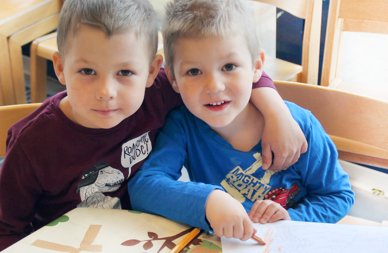 Zwei kleine Jungen sitzen nah beieinander an einem Tisch, der eine hat den Arm um den anderen gelegt und lächelt in die Kamera, während der andere mit einem Buntstift malt.