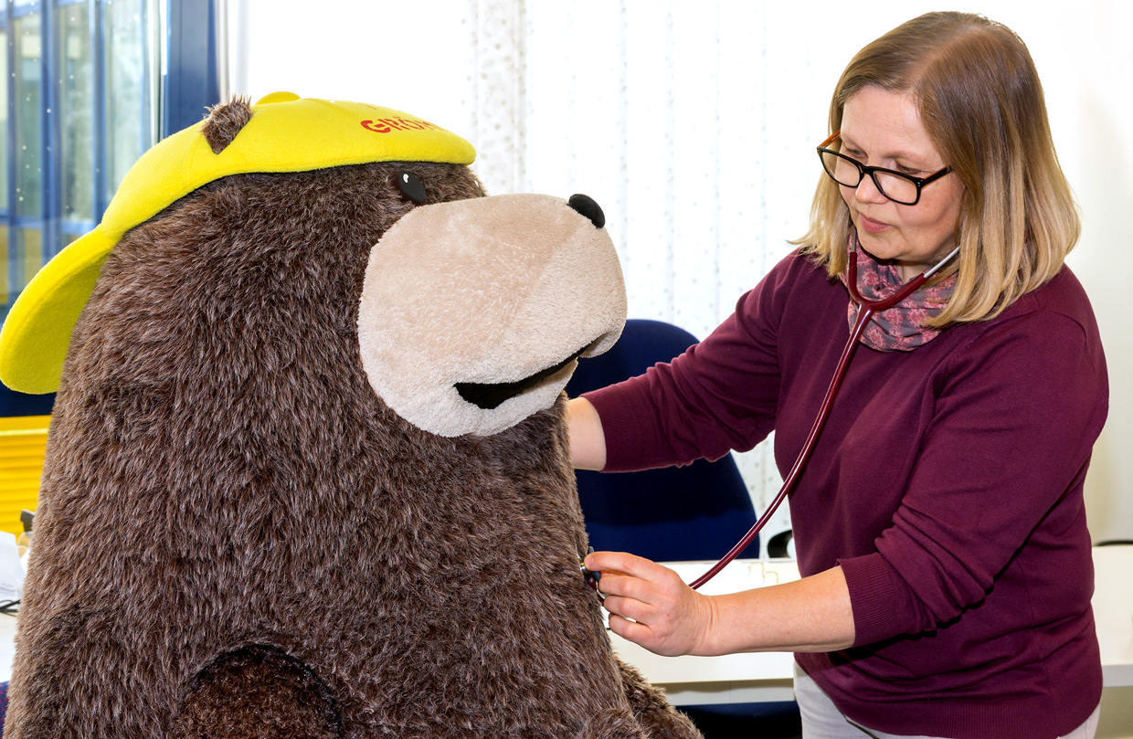Eine Ärztin mit Brille untersucht Grömel, das große, braune Stofftier-Maskottchen des Seeheilbades Grömitz. Grömel trägt eine gelbe Kappe, die Ärztin hält ein Stethoskop an seine Brust.