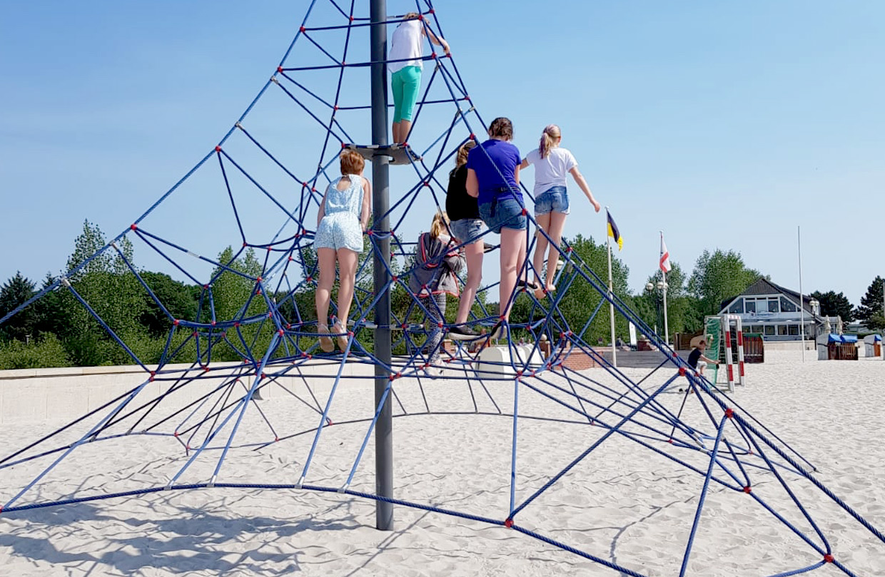 Mehrere Kinder klettern auf einem großen, blauen Seilklettergerüst am Sandstrand der Ostsee unter einem klaren, blauen Himmel. Im Hintergrund sind Bäume und ein Gebäude zu sehen.
