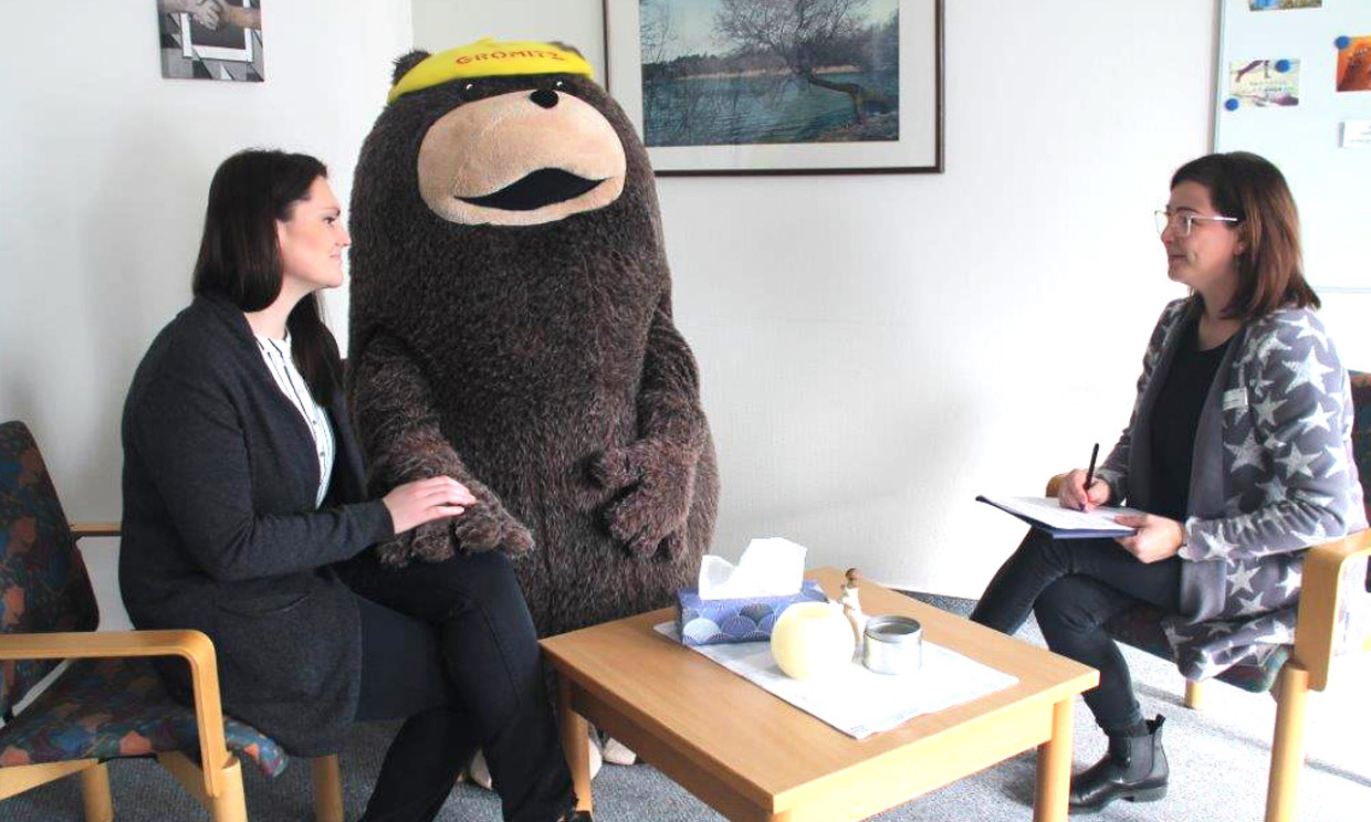 Zwei Frauen, eine Patientin und eine Therapeutin, sitzen sich in einem hellen Raum gegenüber und scheinen sich zu unterhalten. Zwischen ihnen steht das Maskottchen des Seeheilbades Grömitz, ein großer, brauner Bär mit einer gelben Mütze. Die Therapeutin macht Notizen auf einem Klemmbrett.