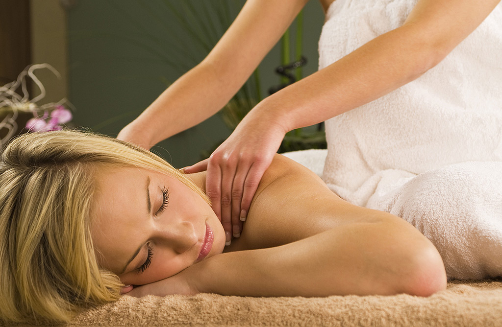 Eine blonde Frau liegt entspannt auf dem Bauch und erhält eine Rückenmassage von den Händen einer Masseurin. Die Frau ist in ein weißes Tuch gehüllt. Der Hintergrund ist unscharf und zeigt grüne und violette Farbtöne.