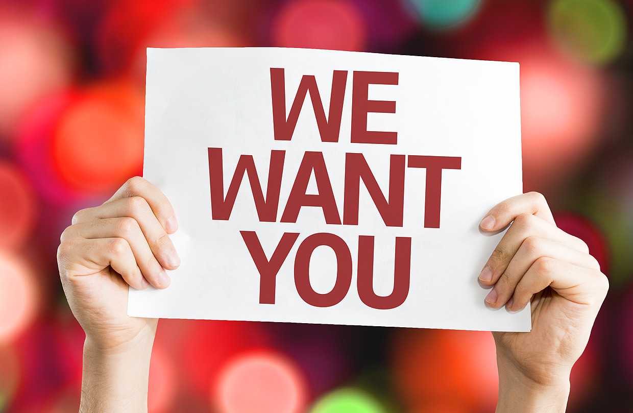 Ein Aufruf für die Seite mit Stellenangeboten der Ostseeklinik Grömitz: Zwei Hände halten ein weißes Schild mit dem rot geschriebenen Text „WE WANT YOU“ vor einem unscharfen Hintergrund mit roten, grünen und weißen Lichtpunkten.