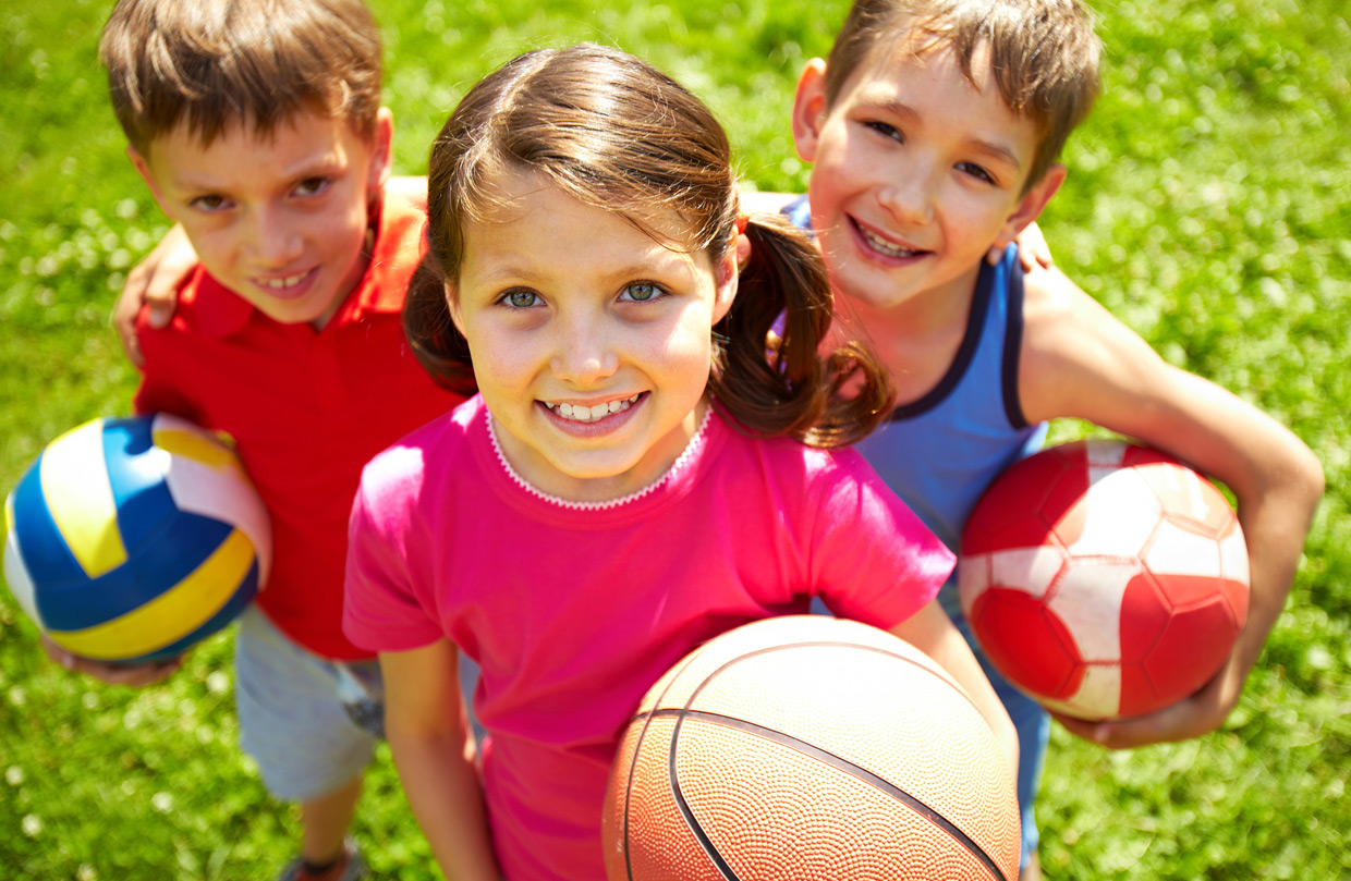 Drei Kinder, ein Mädchen und zwei Jungen, stehen auf einer Wiese und lächeln in die Kamera. Das Mädchen hält einen Basketball in der Hand, die Jungen halten farbige Fußbälle in der Hand und unter dem Arm.