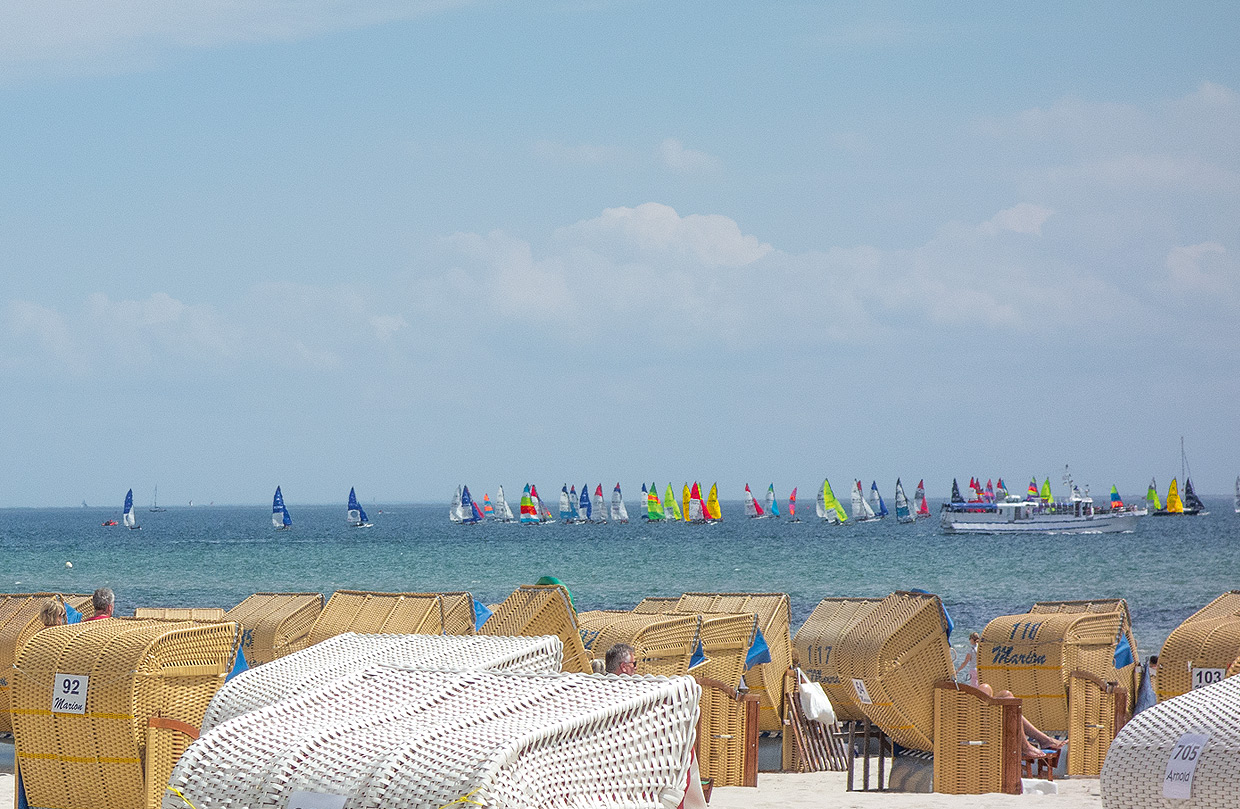 Nahe der Eltern-Kind-Klinik Ostseeklinik Grömitz stehen zahlreiche Strandkörbe am sandigen Ostseestrand von Grömitz. Dahinter erstreckt sich das ruhige, türkisfarbene Meer, auf dem viele kleine Segelboote mit bunten Segeln zu sehen sind. Ein größeres weißes Boot fährt ebenfalls auf dem Wasser. Der Himmel ist hellblau mit vereinzelten weißen Wolken.