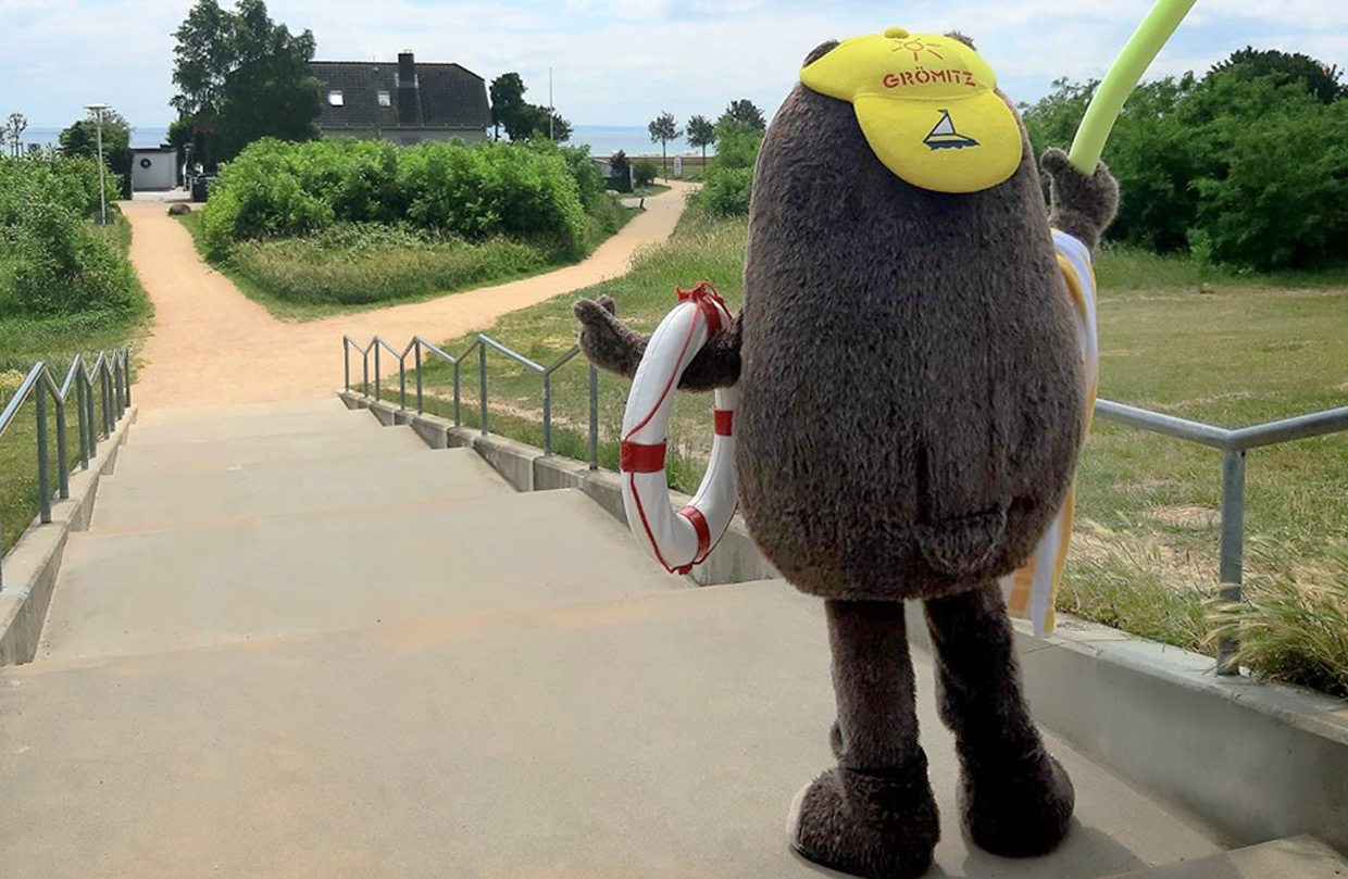 Grömel, das Maskottchen von Grömitz und der Ostseeklinik, als menschengroßes Stofftier mit braunem Fell, gelber Mütze und Rettungsring steht auf einer Treppe, die zu einem Strandweg führt. Im Hintergrund sind Bäume, ein Gebäude und das Meer zu sehen.