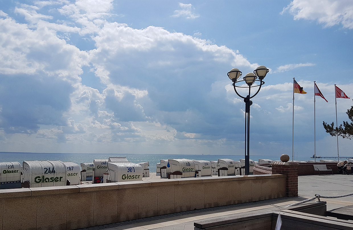 Eine Reihe von Strandkörben steht auf der Strandpromenade vom Grömitz mit Blick auf das Meer unter einem bewölkten Himmel. Im Vordergrund sind eine Laterne und Flaggen zu sehen.