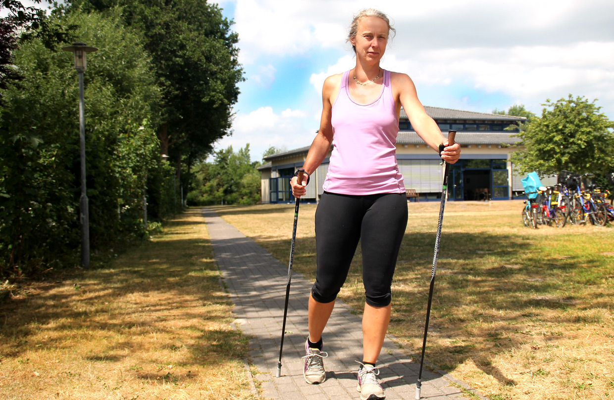 Eine Mutter/Patientin der Ostseeklinik Grömitz in Sportkleidung walkt mit Nordic-Walking-Stöcken im Freien. Im Hintergrund sind Bäume, und ein Klinikgebäude der Eltern-Kind-Kur-Klinik sowie Fahrradständer zu sehen.