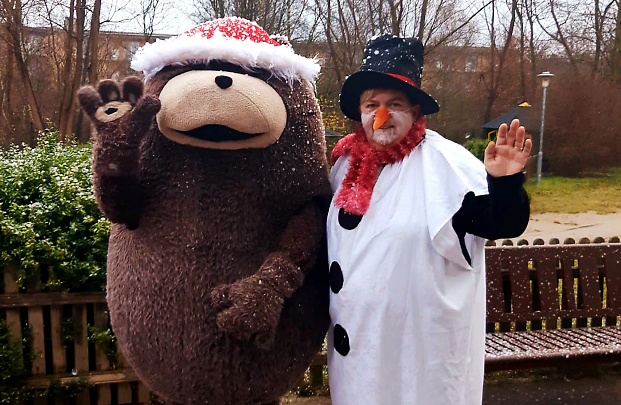 Grömel, das große braune Stofftier-Maskottchen, mit Weihnachtsmütze und ein als Schneemann verkleidete Frau winken gemeinsam in die Kamera im Außenbereich der Klinik. Leichter Schneefall im Hintergrund.