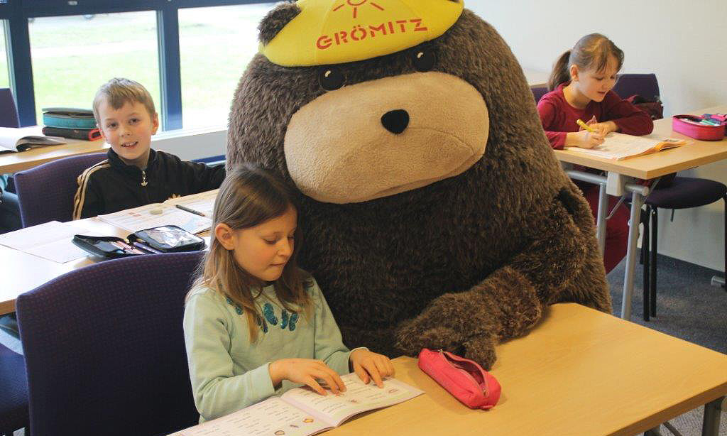 In einem Klassenzimmer steht Grömel, das große Stofftiermaskottchen, neben einem Mädchen an einem Tisch. Das Mädchen liest in einem Schulbuch. Im Hintergrund sind weitere Kinder zu sehen.