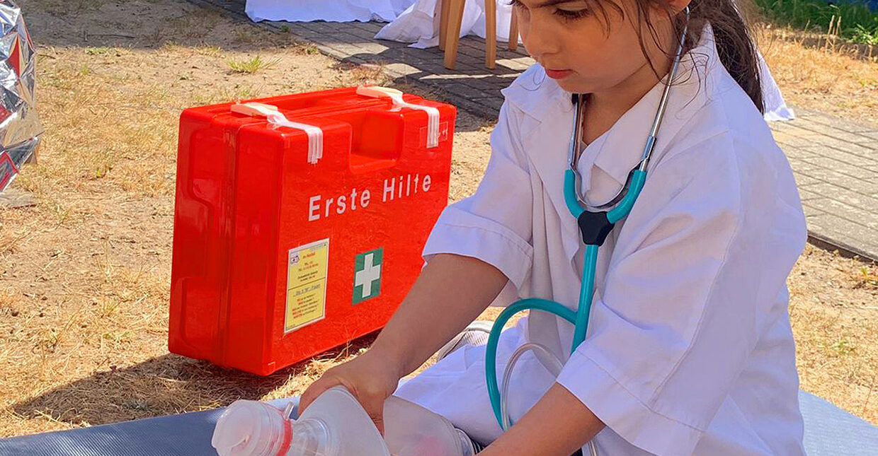 Ein Kind in weißer Kleidung mit einem Stethoskop, das knieend eine Erste-Hilfe-Übung macht. Im Hintergrund ist eine Notfallkoffer zu sehen.